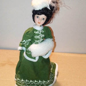 Könnte beinhalten: Eine Porzellanpuppe, die ein grünes Kleid mit weißen Verzierungen und einen weißen Hut mit Federn trägt. Die Puppe hat schwarze Haare und bemalte Gesichtszüge.