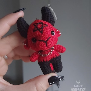 Puede incluir: Una pequeña figura de ganchillo roja y negra con cuernos, un pentagrama bordado en la frente y piernas negras. La figura tiene ojos de plástico negro, pendientes de aro de metal plateado y una cadena plateada alrededor del cuello.