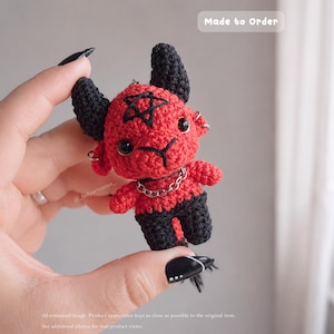 May include: A small crocheted plush toy featuring a red body with black horns, black legs, and a black tail. The figure has black eyes, a stitched pentagram on its forehead, and a silver-colored chain necklace. The text Made to Order is visible.