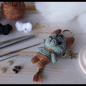 May include: A handmade crochet keychain featuring a small, green creature with brown accents and a tassel tail. The image also shows yarn, scissors, a crochet hook, and other crafting supplies, suggesting a DIY project.