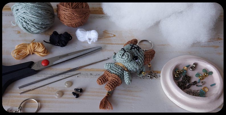 May include: A handmade crochet keychain featuring a small, green creature with brown accents and a tassel tail. The image also shows yarn, scissors, a crochet hook, and other crafting supplies, suggesting a DIY project.