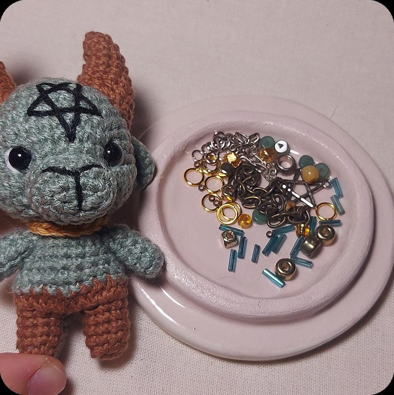 May include: A crocheted teal goat doll with brown horns and legs, a gold collar, and a pentagram on its head. A small pink dish holds various beads, chains, and jewelry findings in gold, silver, and teal.