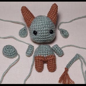 May include: A disassembled crochet bat doll in shades of gray and brown. The bat's head, body, ears, wings, and tail are separated, with long gray yarn strands attached. The bat has black eyes.