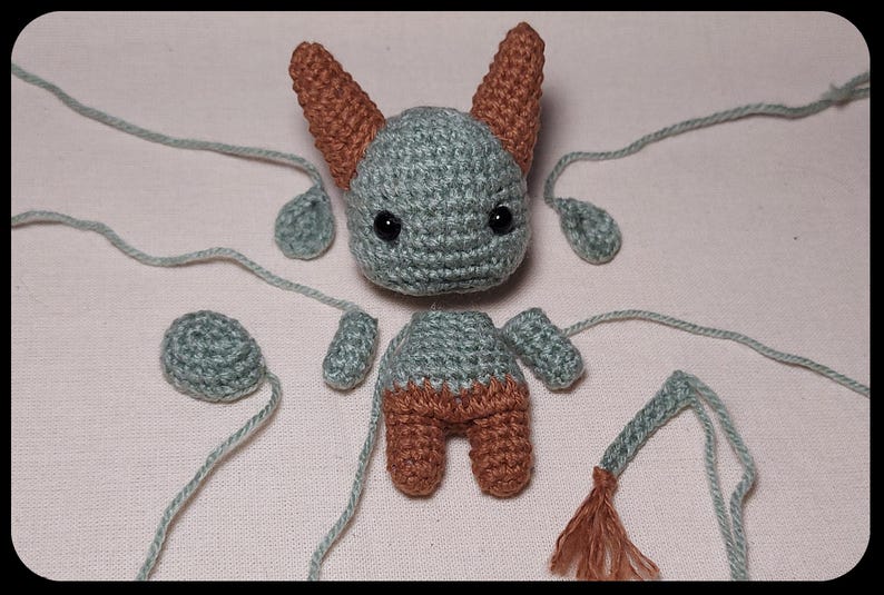 May include: A disassembled crochet bat doll in shades of gray and brown. The bat's head, body, ears, wings, and tail are separated, with long gray yarn strands attached. The bat has black eyes.