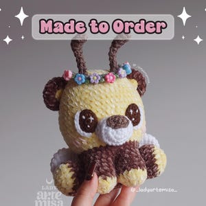 Puede incluir: Un peluche de ciervo tejido a mano, amarillo y marrón, con hocico y patas blancas. Tiene una corona de flores y el texto "Made to Order" encima.