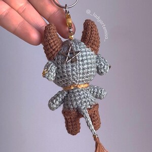 May include: Crocheted keychain featuring a gray and brown horned creature with a star pendant. The handmade charm has a tail with a tassel and a gold-colored keyring. The keychain is a unique accessory.