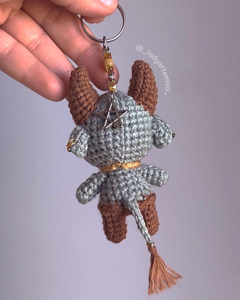 May include: Crocheted keychain featuring a gray and brown horned creature with a star pendant. The handmade charm has a tail with a tassel and a gold-colored keyring. The keychain is a unique accessory.
