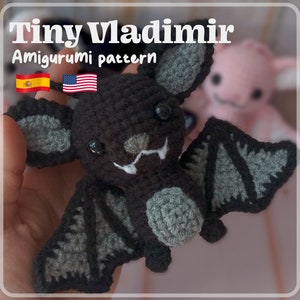 Puede incluir: Un murciélago de amigurumi tejido a crochet con pelaje negro y gris, grandes ojos negros y una sonrisa blanca. El murciélago está siendo sostenido en una mano. La imagen incluye el texto "Tiny Vladimir Amigurumi pattern" y banderas de España y Estados Unidos.