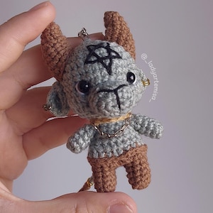 May include: Crocheted gray and brown figurine of a horned creature with a pentagram on its head. The handmade doll has a chain necklace and small earrings. The figurine is held in a hand.