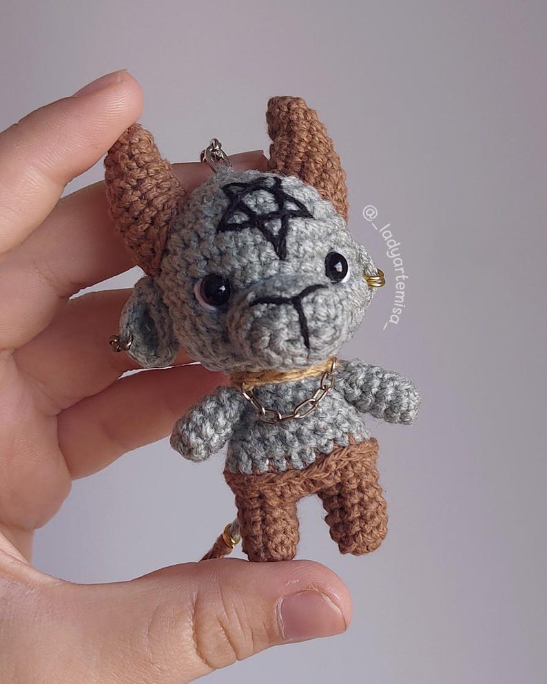May include: Crocheted gray and brown figurine of a horned creature with a pentagram on its head. The handmade doll has a chain necklace and small earrings. The figurine is held in a hand.