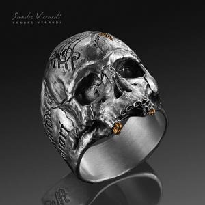Puede incluir: Un anillo de plata con forma de calavera con detalles dorados. La calavera est&aacute; detallada con tallas intrincadas y tiene una superficie texturizada.