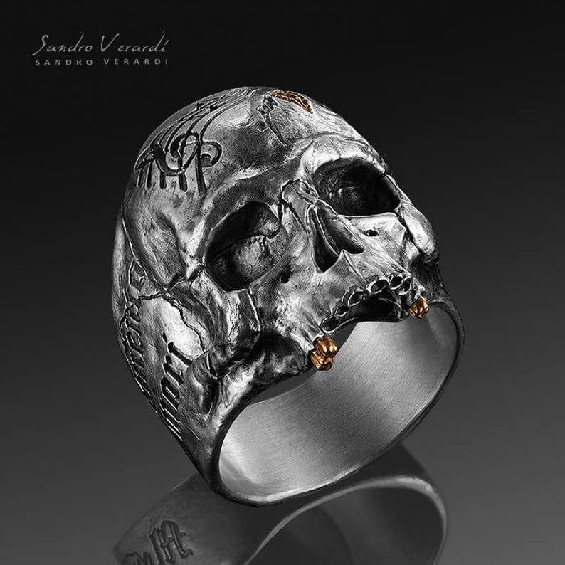 Puede incluir: Un anillo de plata con forma de calavera con detalles dorados. La calavera est&aacute; detallada con tallas intrincadas y tiene una superficie texturizada.