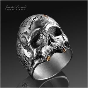 Puede incluir: Un anillo de plata con forma de calavera con detalles dorados. La calavera est&aacute; detallada con una superficie texturizada y las palabras "Invidio", "Superba" y "Accedo" est&aacute;n inscritas en la parte superior de la calavera.