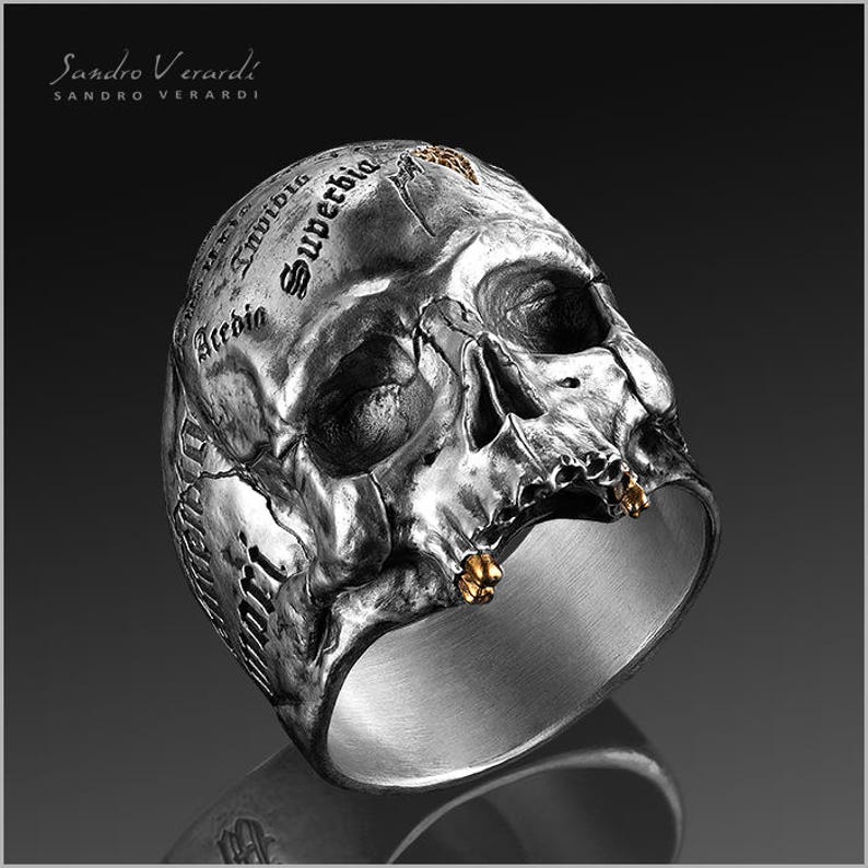 Puede incluir: Un anillo de plata con forma de calavera con detalles dorados. La calavera est&aacute; detallada con una superficie texturizada y las palabras "Invidio", "Superba" y "Accedo" est&aacute;n inscritas en la parte superior de la calavera.