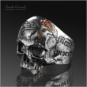 Puede incluir: Un anillo de calavera de plata con una rosa y las palabras "Memento vivere" grabadas en el lateral.