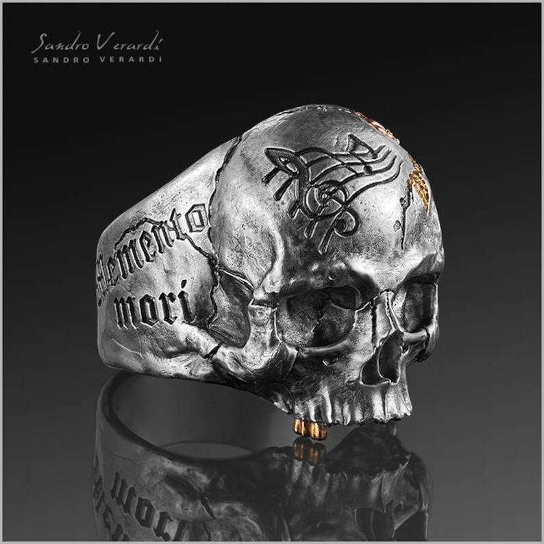 Puede incluir: Un anillo de calavera de plata con un dise&ntilde;o detallado y la inscripci&oacute;n "Memento Mori" en el lateral. El anillo tiene un acento dorado en la parte superior.