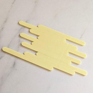 May include: A set of 10 yellow wooden craft sticks, arranged in a staggered pattern on a white marble surface.