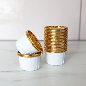 Peut inclure: Un lot de caissettes à cupcakes en papier blanc avec des bords dorés. Les caissettes sont empilées sur une surface en marbre blanc.