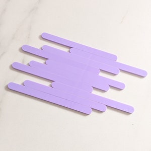 May include: A set of ten lavender colored wooden craft sticks. The sticks are arranged in a staggered pattern on a white marble surface.
