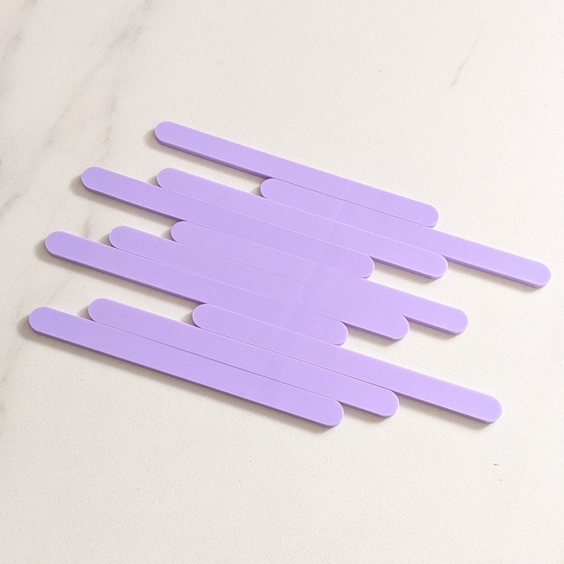 May include: A set of ten lavender colored wooden craft sticks. The sticks are arranged in a staggered pattern on a white marble surface.