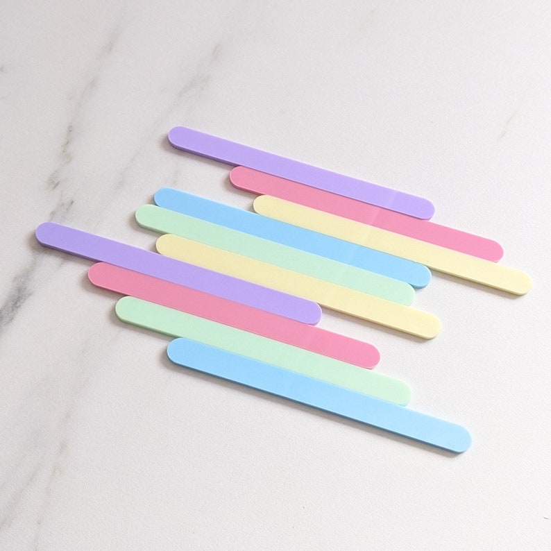 May include: A set of eight pastel-colored wooden craft sticks arranged in a diagonal pattern on a white marble surface. The sticks are in shades of light blue, green, pink, yellow, and purple.