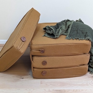 Peut inclure: Quatre coussins carrés de couleur moutarde avec des coins arrondis et un bouton marron sur le côté de chacun. Un coussin est posé verticalement tandis que trois sont empilés horizontalement. Un tissu frangé vert olive foncé est drapé.