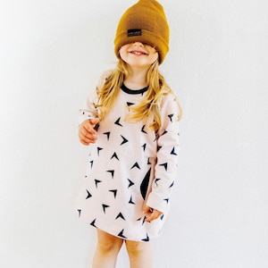 Pode incluir: Uma pessoa jovem vestindo um vestido rosa e preto com estampa geométrica de mangas compridas. Ela está usando um gorro amarelo mostarda sobre os olhos.