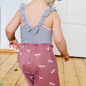 Op de afbeelding: Een kinderoutfit met een blauw-wit gestreept body met roze rand en strik aan de achterkant. De mauvekleurige shorts zijn versierd met witte libellenpatronen. Een lichtblauwe zonnehoed maakt de set compleet.