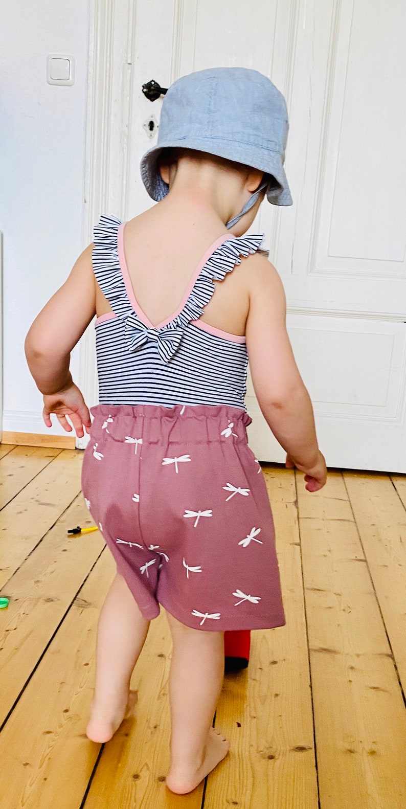 Op de afbeelding: Een kinderoutfit met een blauw-wit gestreept body met roze rand en strik aan de achterkant. De mauvekleurige shorts zijn versierd met witte libellenpatronen. Een lichtblauwe zonnehoed maakt de set compleet.