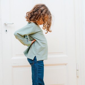 Puede incluir: Una persona joven con una camisa de manga larga verde claro y pantalones vaqueros azules. La persona est&aacute; de pie frente a una puerta blanca.