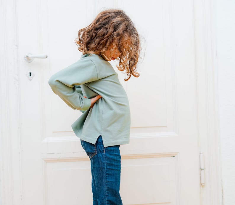 Puede incluir: Una persona joven con una camisa de manga larga verde claro y pantalones vaqueros azules. La persona est&aacute; de pie frente a una puerta blanca.
