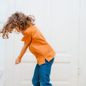 Puede incluir: Una persona joven con cabello casta&ntilde;o rizado lleva una camiseta de color &oacute;xido y pantalones vaqueros azules bailando frente a una puerta blanca.