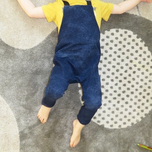 May include: A child wearing a blue denim overall jumpsuit with yellow straps over a yellow short-sleeved shirt. The child is lying on a gray and white patterned rug.
