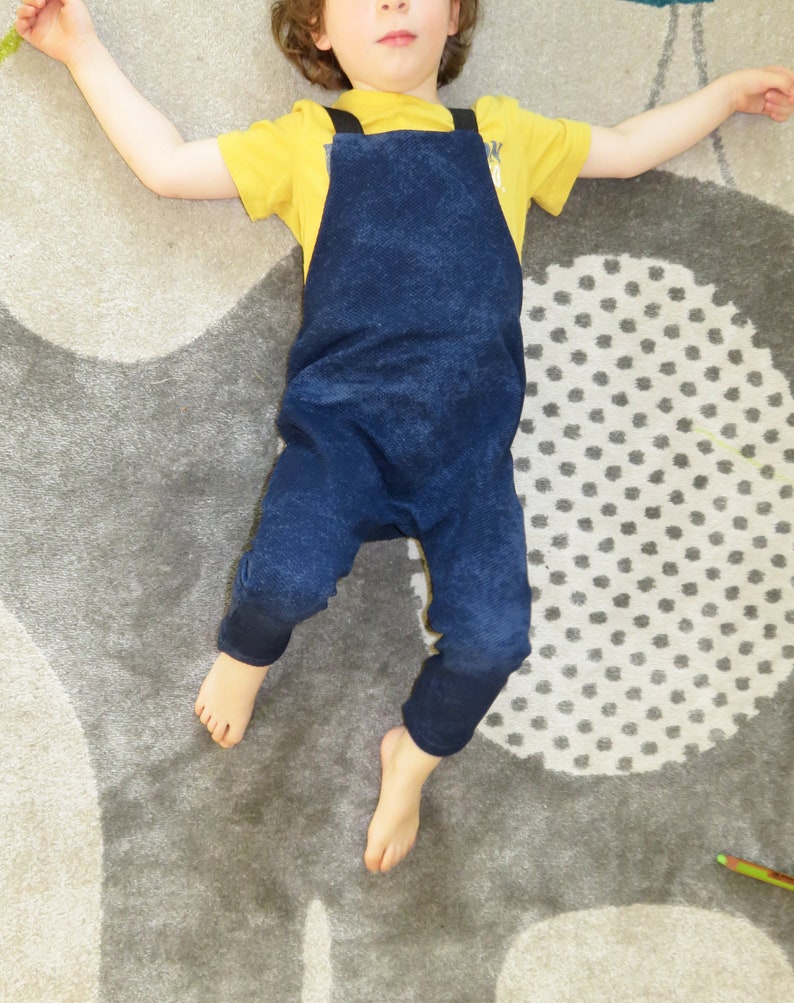 May include: A child wearing a blue denim overall jumpsuit with yellow straps over a yellow short-sleeved shirt. The child is lying on a gray and white patterned rug.