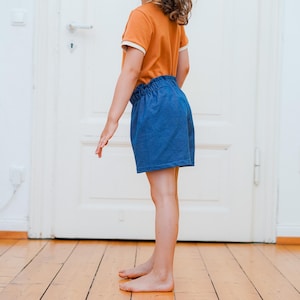 Op de afbeelding: Een kind draagt een oranje t-shirt met witte bies en een blauwe korte broek. De korte broek heeft een elastische tailleband. Het kind staat op een houten vloer voor een witte deur.