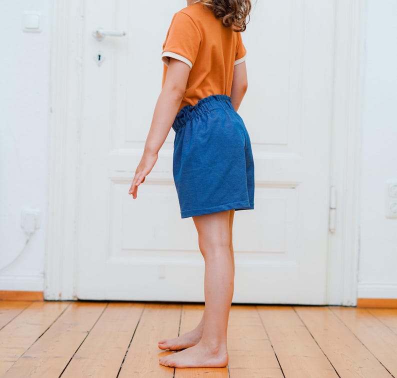 Op de afbeelding: Een kind draagt een oranje t-shirt met witte bies en een blauwe korte broek. De korte broek heeft een elastische tailleband. Het kind staat op een houten vloer voor een witte deur.