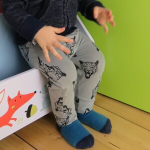 May include: A child wearing gray pants with a black and white animal print. The pants have a tiger face design. The child is sitting on a white and blue toy box.