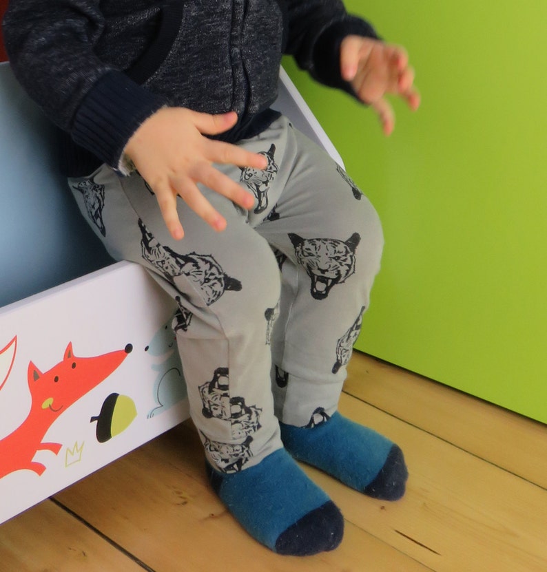 May include: A child wearing gray pants with a black and white animal print. The pants have a tiger face design. The child is sitting on a white and blue toy box.