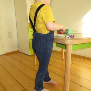 May include: A young child wearing a yellow shirt and blue pants with black suspenders stands near a wooden table with a green top. The child is barefoot and looking down at something on the table.