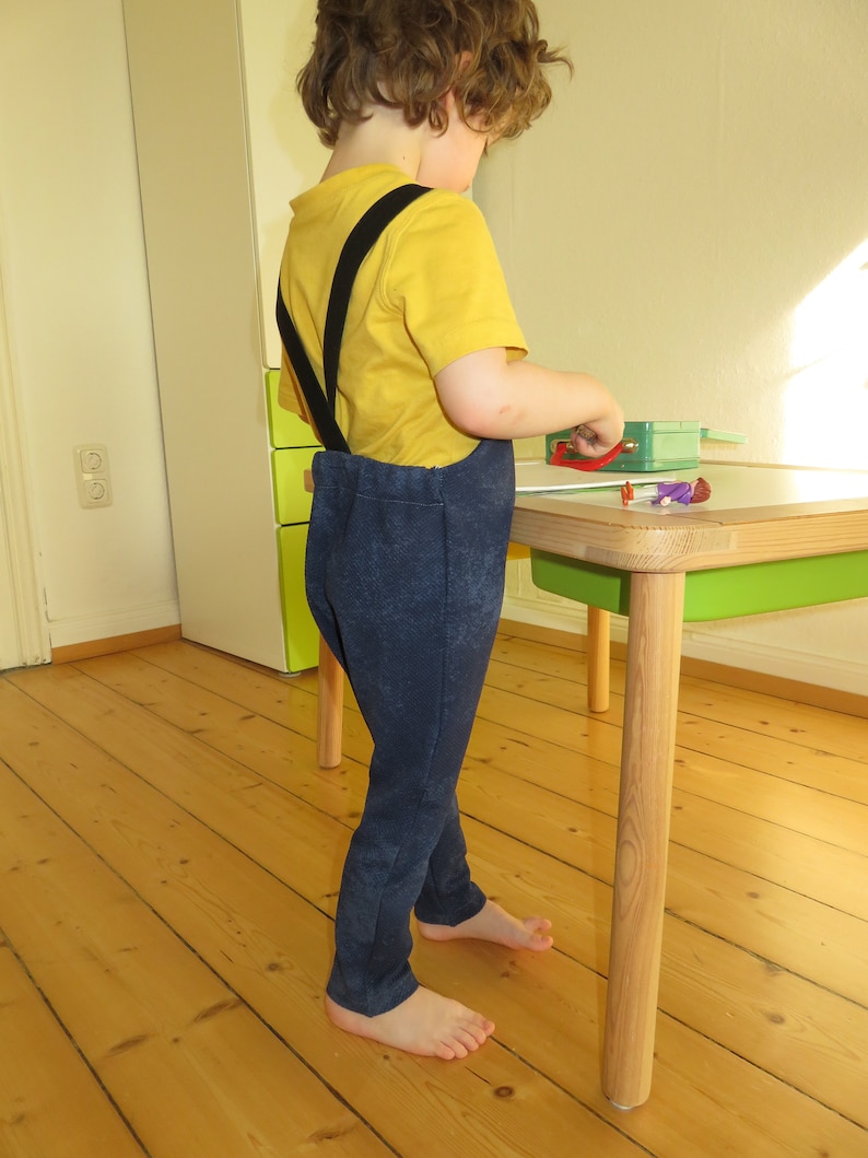 May include: A young child wearing a yellow shirt and blue pants with black suspenders stands near a wooden table with a green top. The child is barefoot and looking down at something on the table.