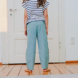 May include: A person wearing a striped short-sleeve shirt and light blue pants. The shirt has horizontal navy blue stripes on a white background. The pants are a solid light blue color and have a wide-leg design. The person is standing in front of a white door.