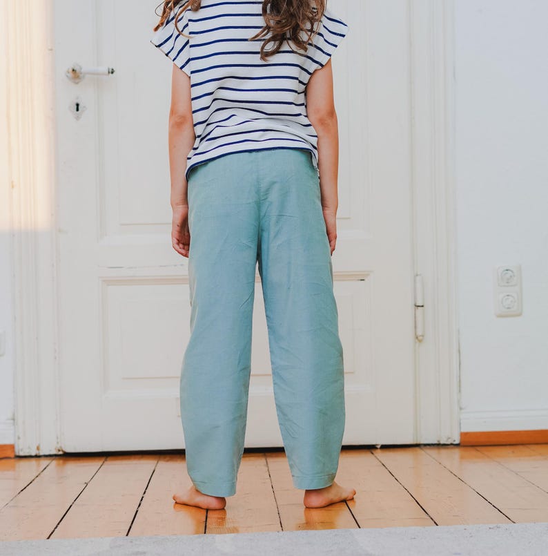 May include: A person wearing a striped short-sleeve shirt and light blue pants. The shirt has horizontal navy blue stripes on a white background. The pants are a solid light blue color and have a wide-leg design. The person is standing in front of a white door.