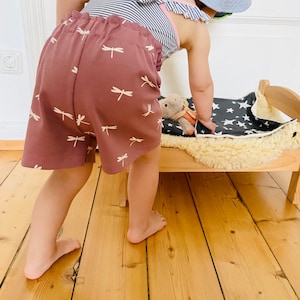 Op de afbeelding: Een kind draagt een gestreepte zwemkleding met ruches en mauve shorts met witte libellenpatronen. Het kind buigt zich over een klein houten bed met een sterrenpatroon deken en een knuffel.