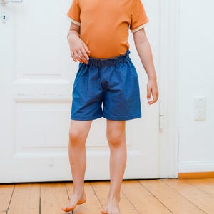 Op de afbeelding: Een kind draagt een oranje t-shirt met witte rand en een blauwe spijkerbroek. De shorts hebben een elastische tailleband. Het kind staat op een houten vloer voor een witte deur.