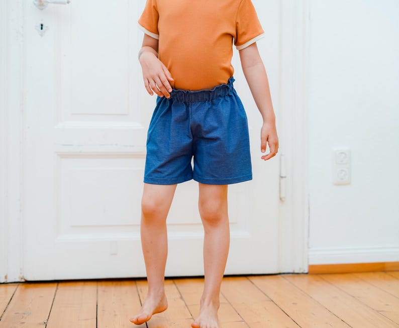Op de afbeelding: Een kind draagt een oranje t-shirt met witte rand en een blauwe spijkerbroek. De shorts hebben een elastische tailleband. Het kind staat op een houten vloer voor een witte deur.