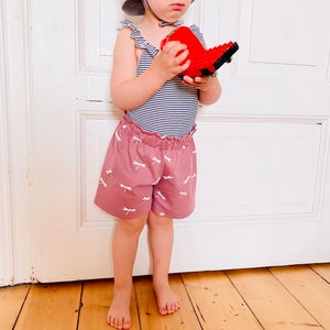 Op de afbeelding: Een kind draagt een gestreepte bodysuit, roze shorts met witte libellenpatronen, een grijze zonnehoed en een groene zonnebril. Het kind houdt een rood speelgoed vast. De outfit is geschikt voor warm weer.