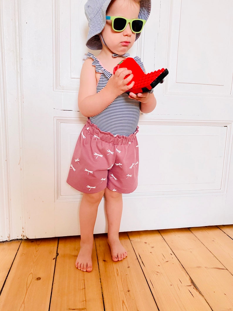 Op de afbeelding: Een kind draagt een gestreepte bodysuit, roze shorts met witte libellenpatronen, een grijze zonnehoed en een groene zonnebril. Het kind houdt een rood speelgoed vast. De outfit is geschikt voor warm weer.
