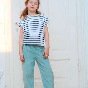 May include: A young person wearing a white and navy blue striped t-shirt and light blue pants. The t-shirt has short sleeves. The pants are loose-fitting. The person is standing in front of a white door.