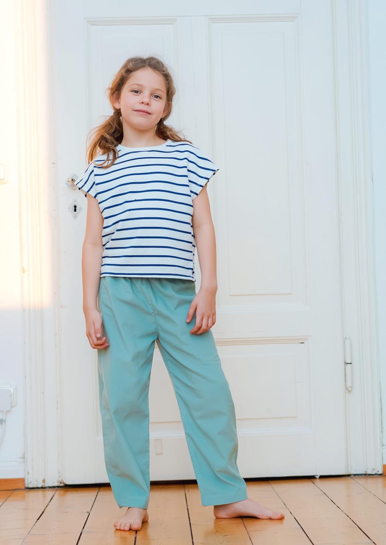May include: A young person wearing a white and navy blue striped t-shirt and light blue pants. The t-shirt has short sleeves. The pants are loose-fitting. The person is standing in front of a white door.