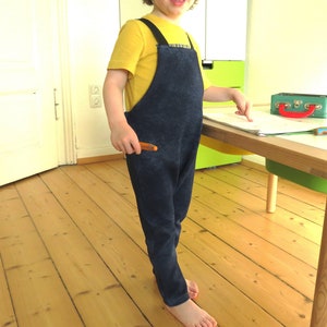 May include: A child wearing a blue denim overall with a yellow shirt and a brown crayon in their hand.